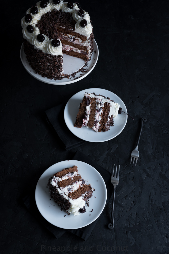 tall chocolate layer cake covered in chocolate curls on a white cake stand, filled and decorated with whipped cream and dark cherries two slices of cake on plates