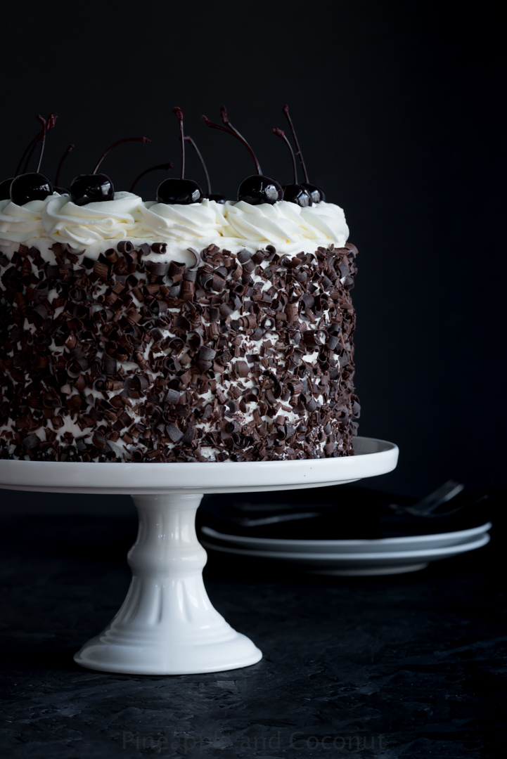 ayer cake covered in chocolate curls on a white cake stand, decorated with whipped cream and dark cherries