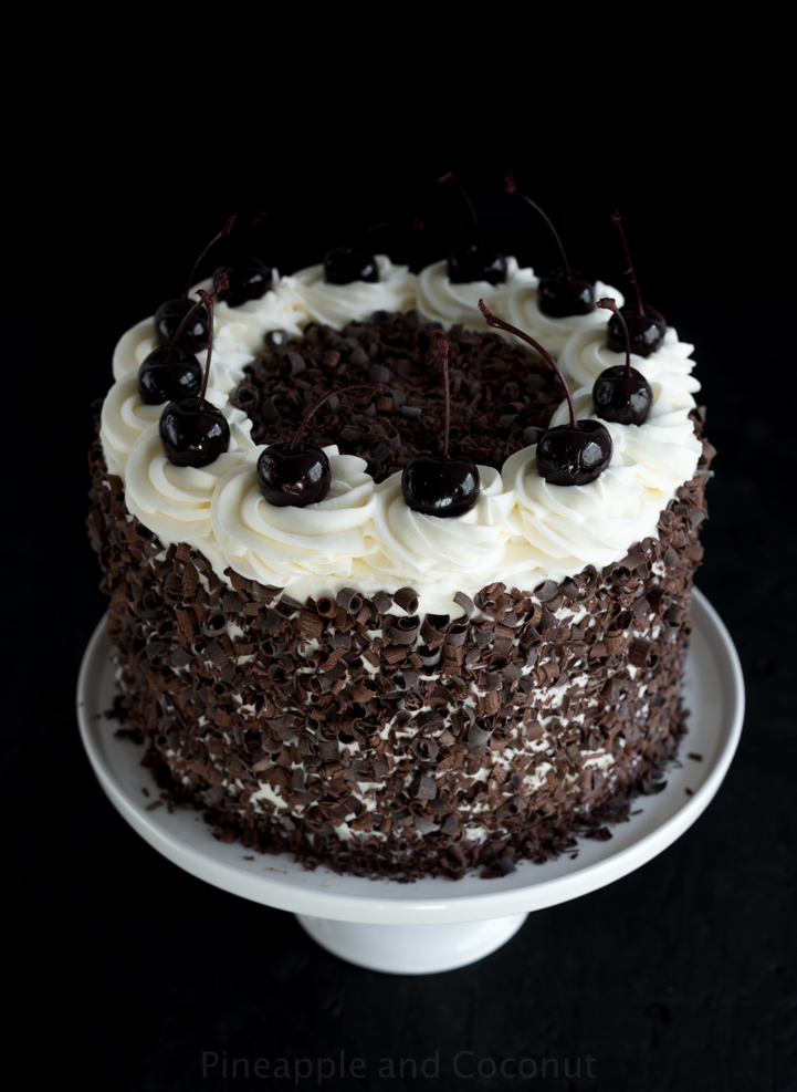 layer cake covered in chocolate curls on a white cake stand, decorated with whipped cream and dark cherries