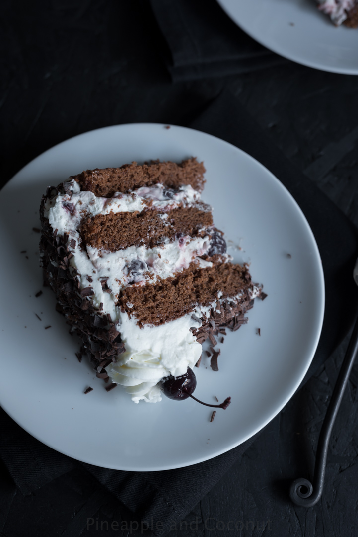 slice of chocolate layer cake on white plate, cake filled with whipped cream and cherries, chocolate curls, fork