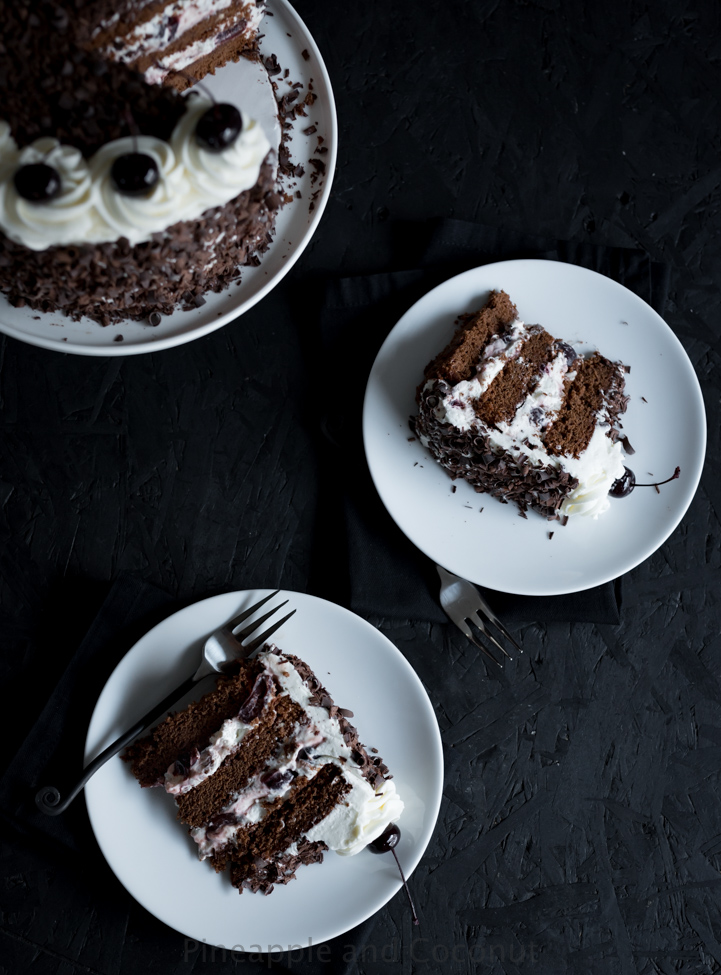 tall chocolate layer cake covered in chocolate curls on a white cake stand, filled and decorated with whipped cream and dark cherries two slices of cake on plates