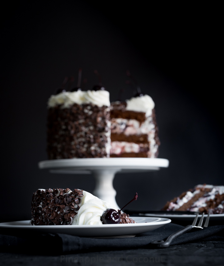 layer cake covered in chocolate curls on a white cake stand, decorated with whipped cream and dark cherries two slices of cake on plates