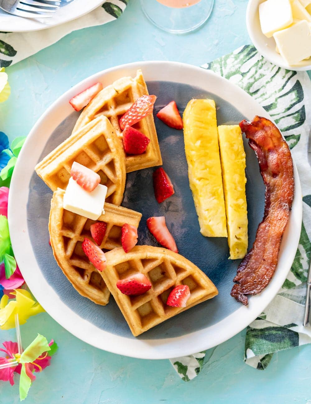 coconut mochi waffles on blue plates with pineapple spears, pieces of cooked bacon, glass with mimosa cocktail in it, glass syrup bottle, rainbow Hawaiian lei on table