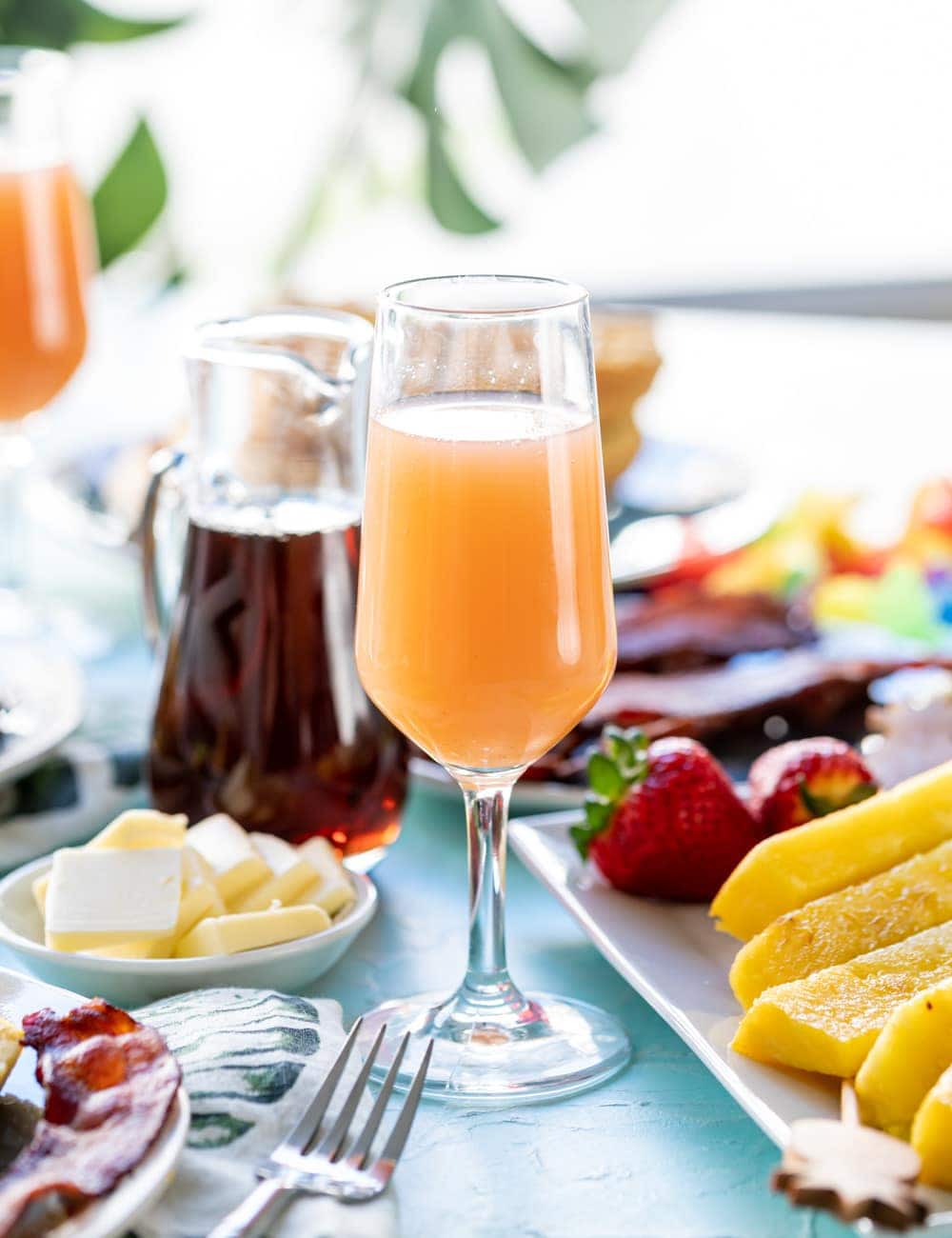 Guava Mimosa in a glass, pineapple spears on plate next to drink, butter on a dish, glass bottle of maple syrup