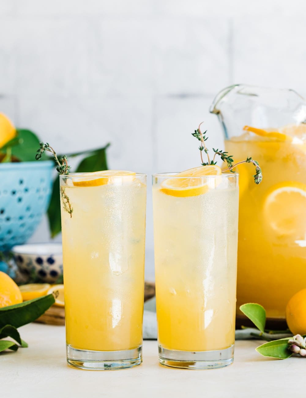 two tall collins glasses filled with lemonade, lemon slices and fresh thyme in glasses, pitcher or lemonade, fresh whole lemons, blue bowl with lemons, cutting board with lemon slices