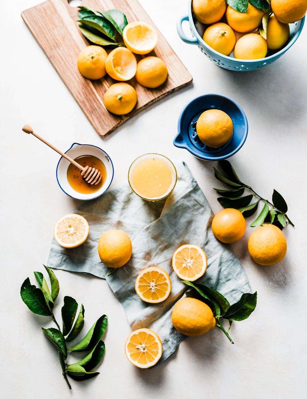 cut and whole lemons, cutting board with lemons, blue bowl filled with lemons, lemon leaves, honey in a small bowl with honey stick, glass of lemon juice