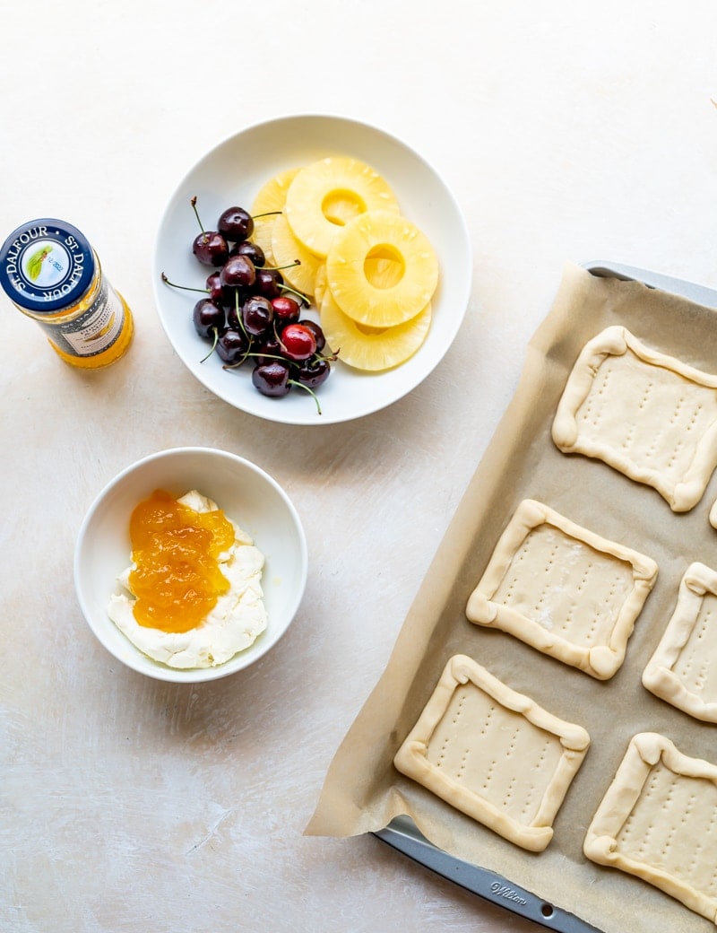 Prep photo for pineapple tarts, cream cheese and jam in small white bowl, cherries and pineapple rings in small white plate, jar of jam, beige background