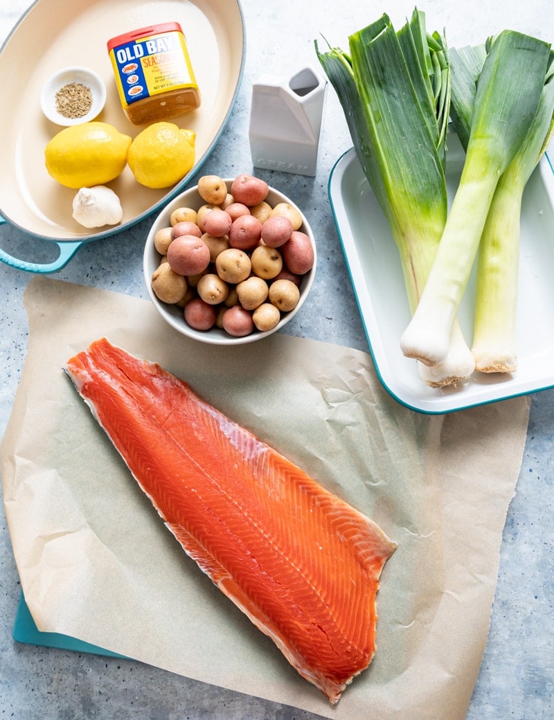 sockeye salmon fillet on paper, tray with three whole leeks, bowl of baby potatoes, aqua pan with lemons, spices, garlic, cream container