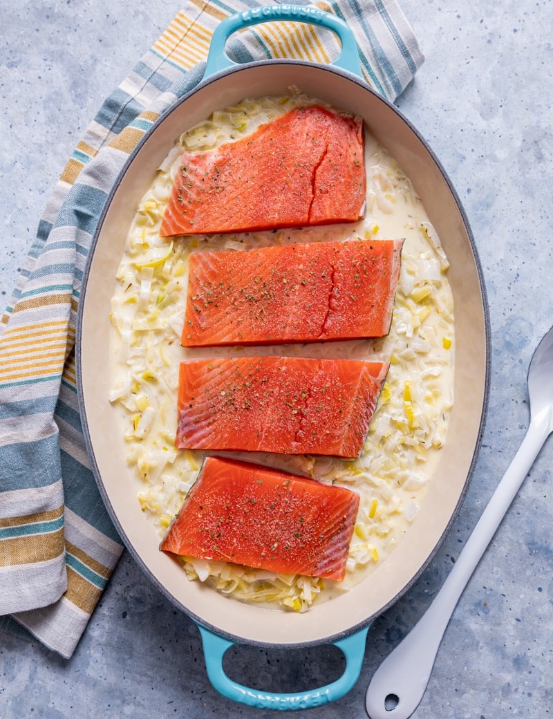 oval aqua cast iron skillet with raw sliced leeks in cream sauce, raw seasoned salmon over the leeks, yellow and blue striped napkin, white spoon