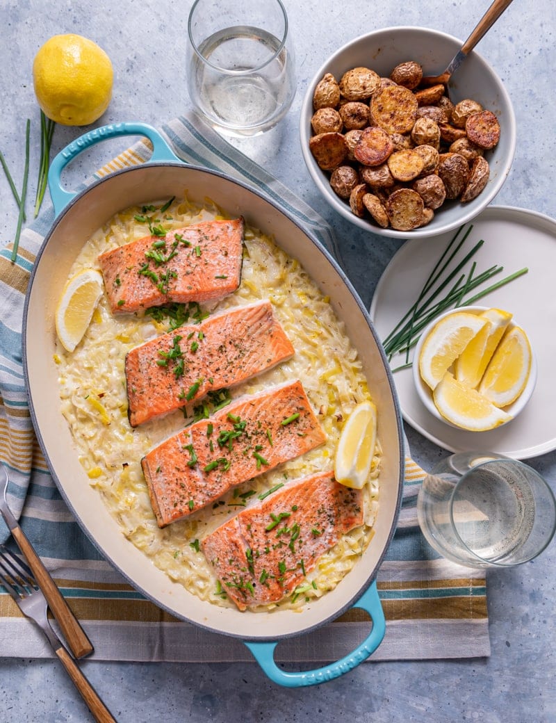 aqua cast iron skilled with baked seasoned salmon on top of leeks with cream sauce, lemon slices, bowl of roasted seasoned baby potatoes