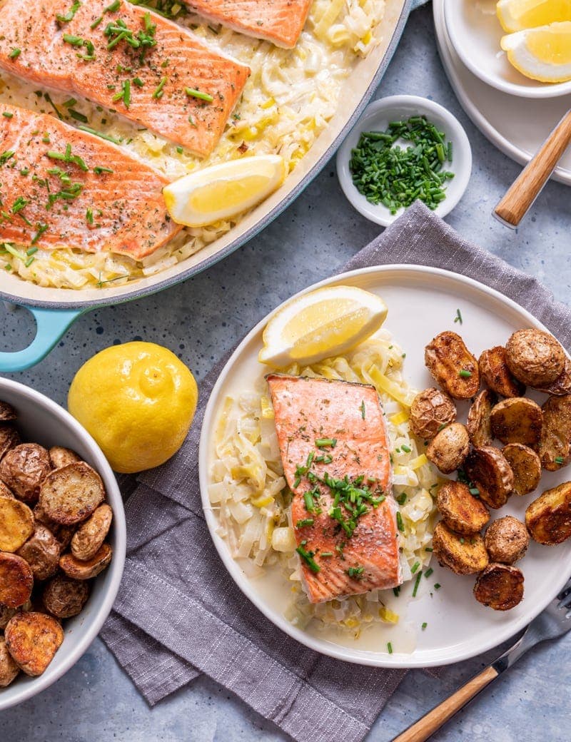 white plate with a piece of salmon over leeks, potatoes on plate as well as a bowl to the side of the dish, whole lemon, lemon slices, large pan of salmon fillets over leeks