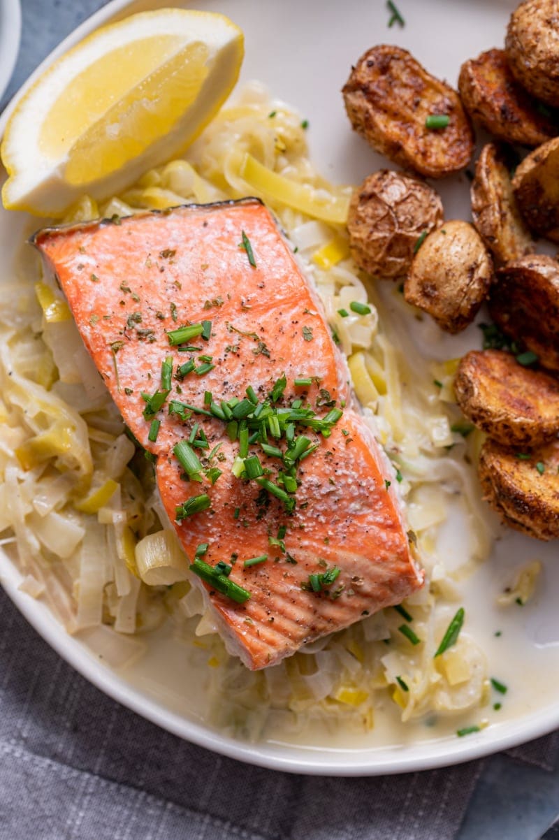 close up of a salmon fillet on top of a serving of cooked creamed leeks, lemon slice, old bay roasted baby potatoes