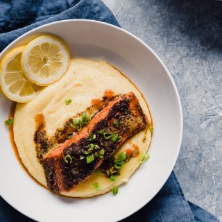 White bowl with yellow grits, seasoned salmon fillet, lemon slices in bowl, gray background, navy blue napkin