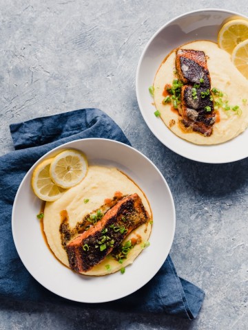 Two white bowls on gray background, with yellow grits, salmon fillets with seasoning, lemon slices and a navy blue napkin