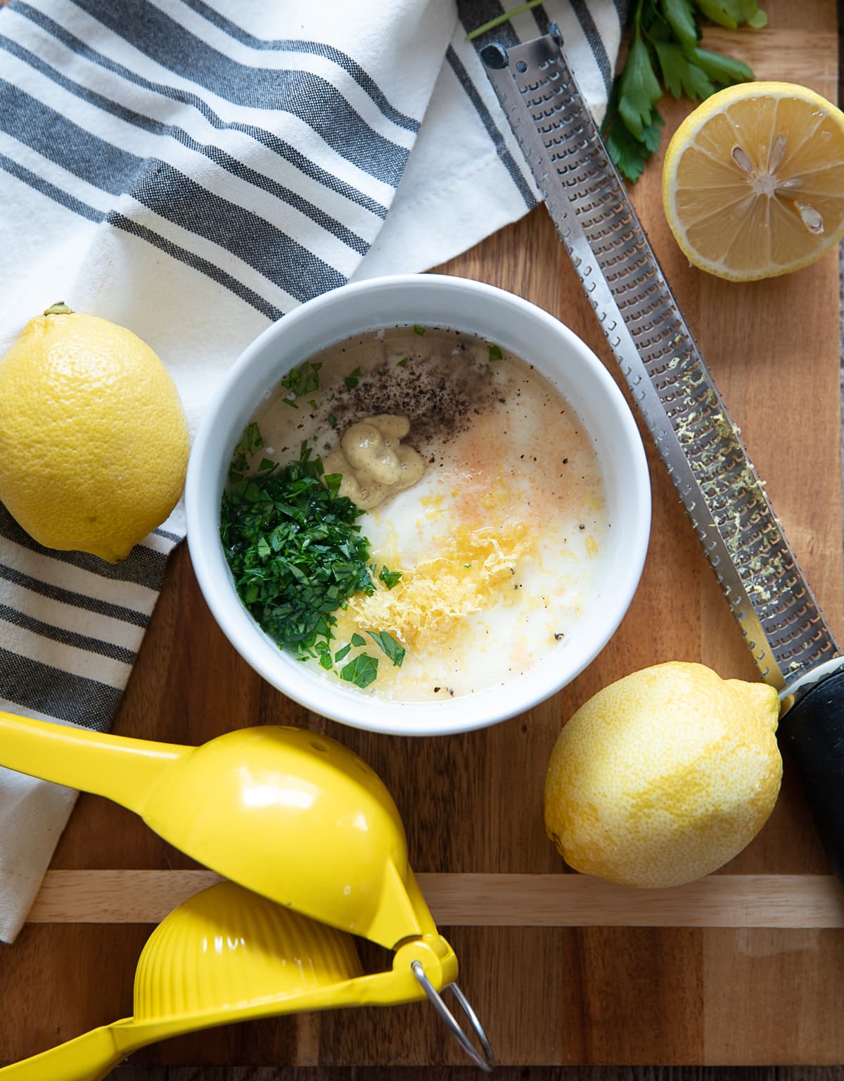 white bowl with lemon mayonnaise parsley, mustard, whole lemons, lemon zester, yellow lemon press, blue and white striped napkin