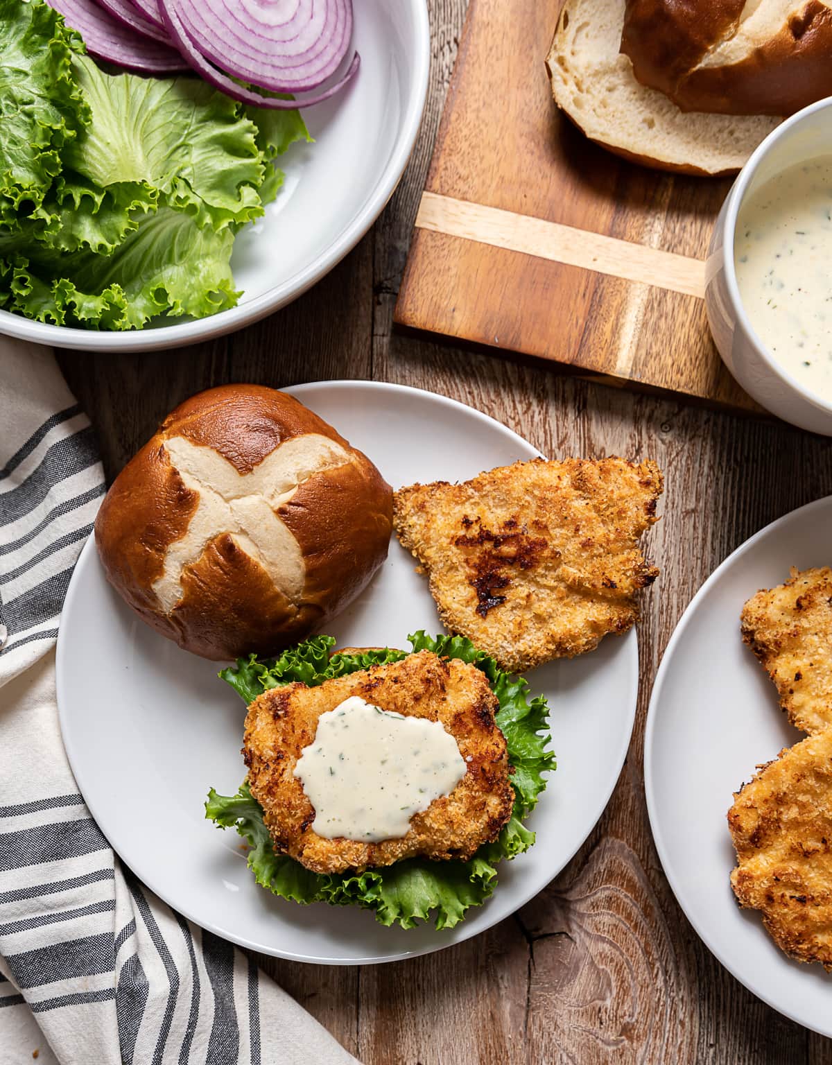 white plate with open faced sandwich , breaded fried chick on top of lettuce, white mayonnaise sauce, red onion rings, plate with thin fried chicken, bottle and glass of beer, bowl with lettuce and onions, cutting board with pretzel buns and bowl of mayonnaise sauce