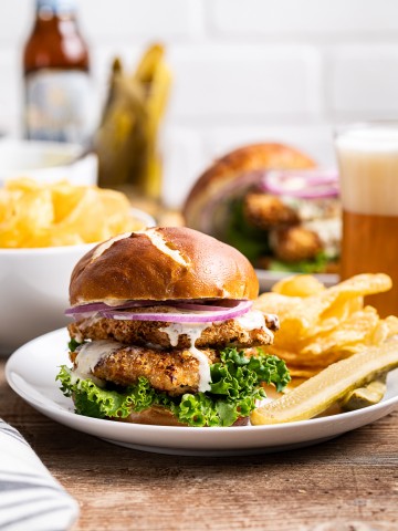 chicken schnitzel sandwiches on pretzel bun with green lettuce red onion kettle chips pickles and glass of beer