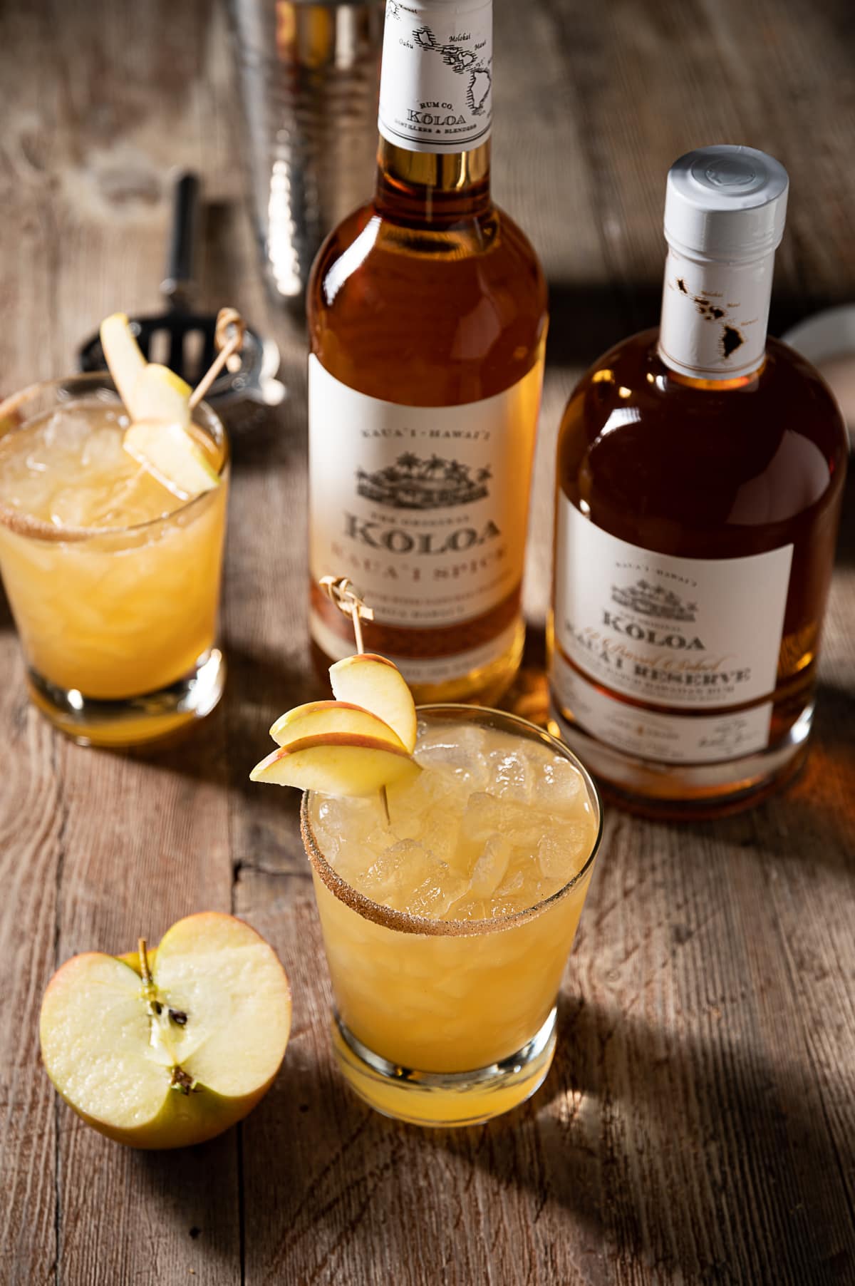 gold colored alcohol mixture in rocks glasses filled with ice, thin apple slices, cocktail shaker, bottles of rum