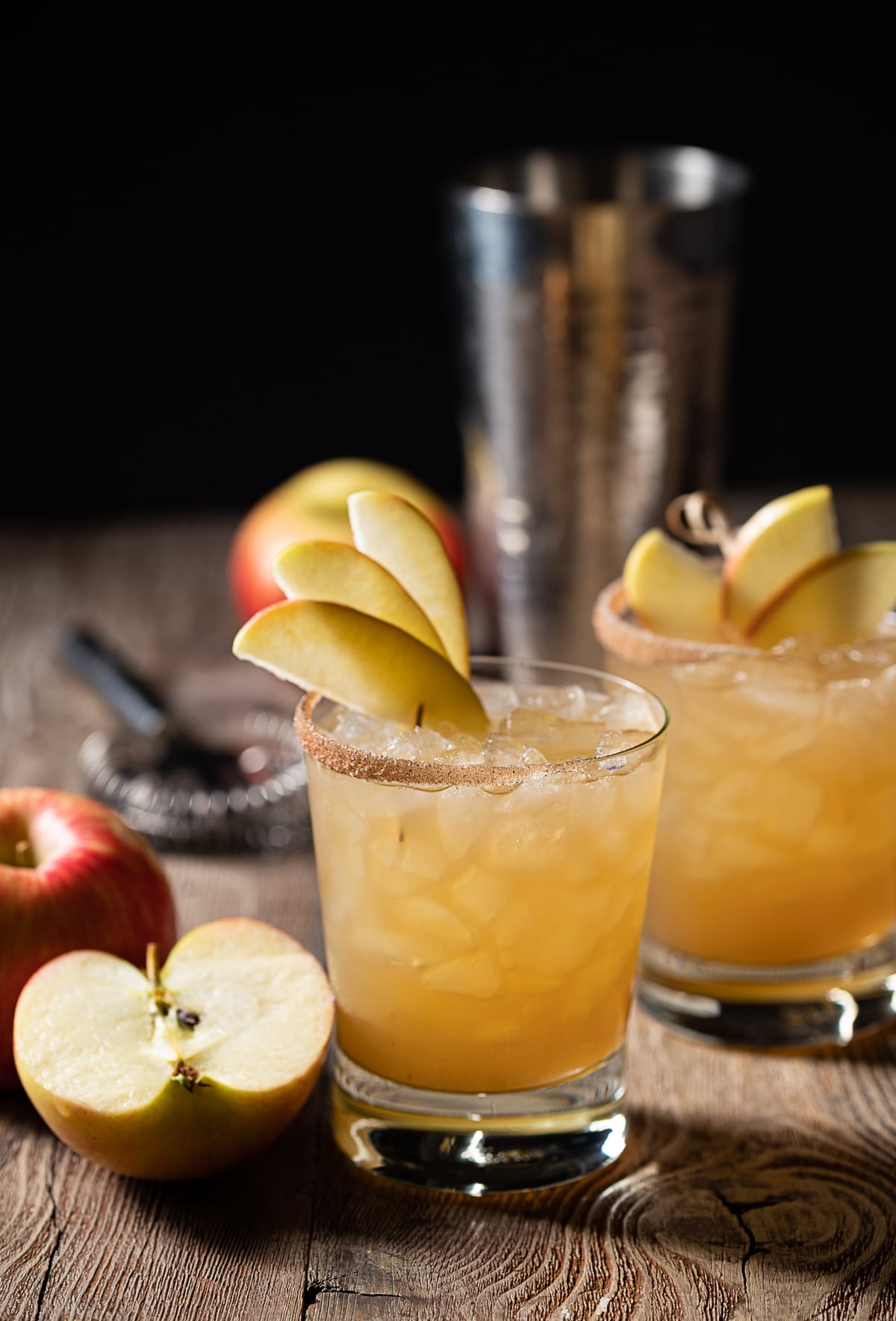 gold colored alcohol mixture in rocks glasses filled with ice, thin apple slices, cocktail shaker, half an apple