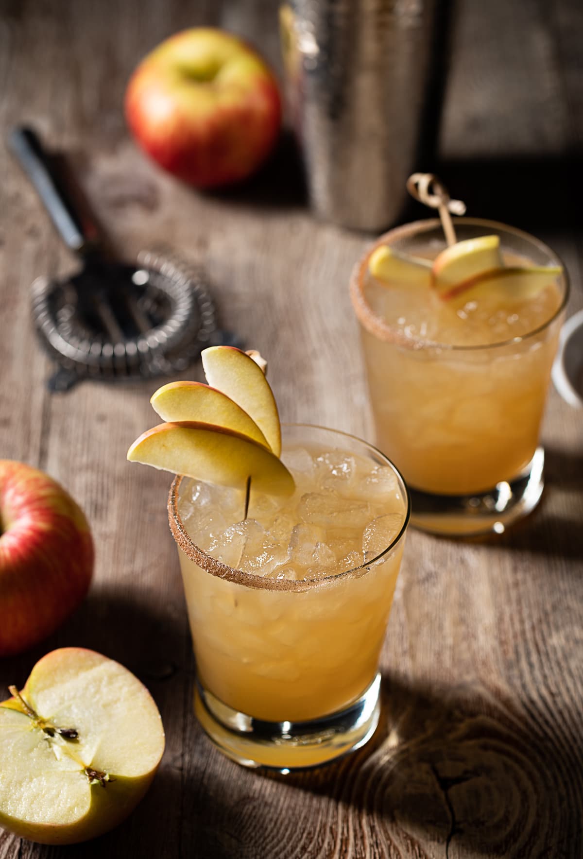 gold colored alcohol mixture in rocks glasses filled with ice, thin apple slices, cocktail shaker, half an apple, two whole apples