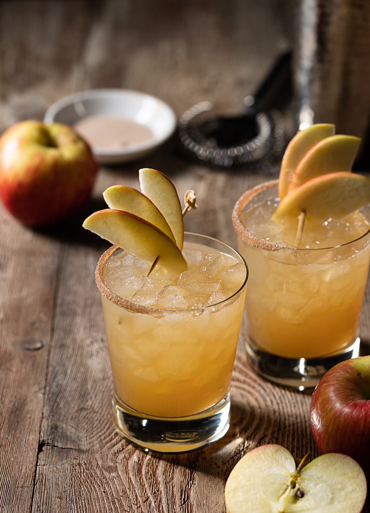 gold colored alcohol mixture in rocks glasses filled with ice, thin apple slices, cocktail shaker, half an apple