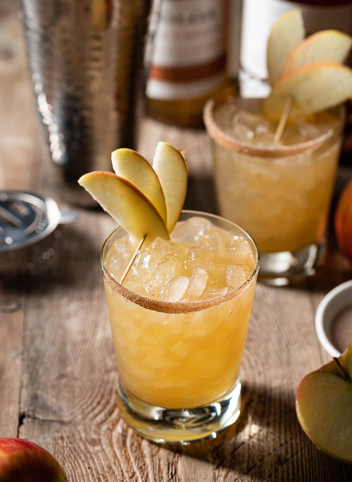 gold colored alcohol mixture in rocks glasses filled with ice, thin apple slices, cocktail shaker, bottles of rum