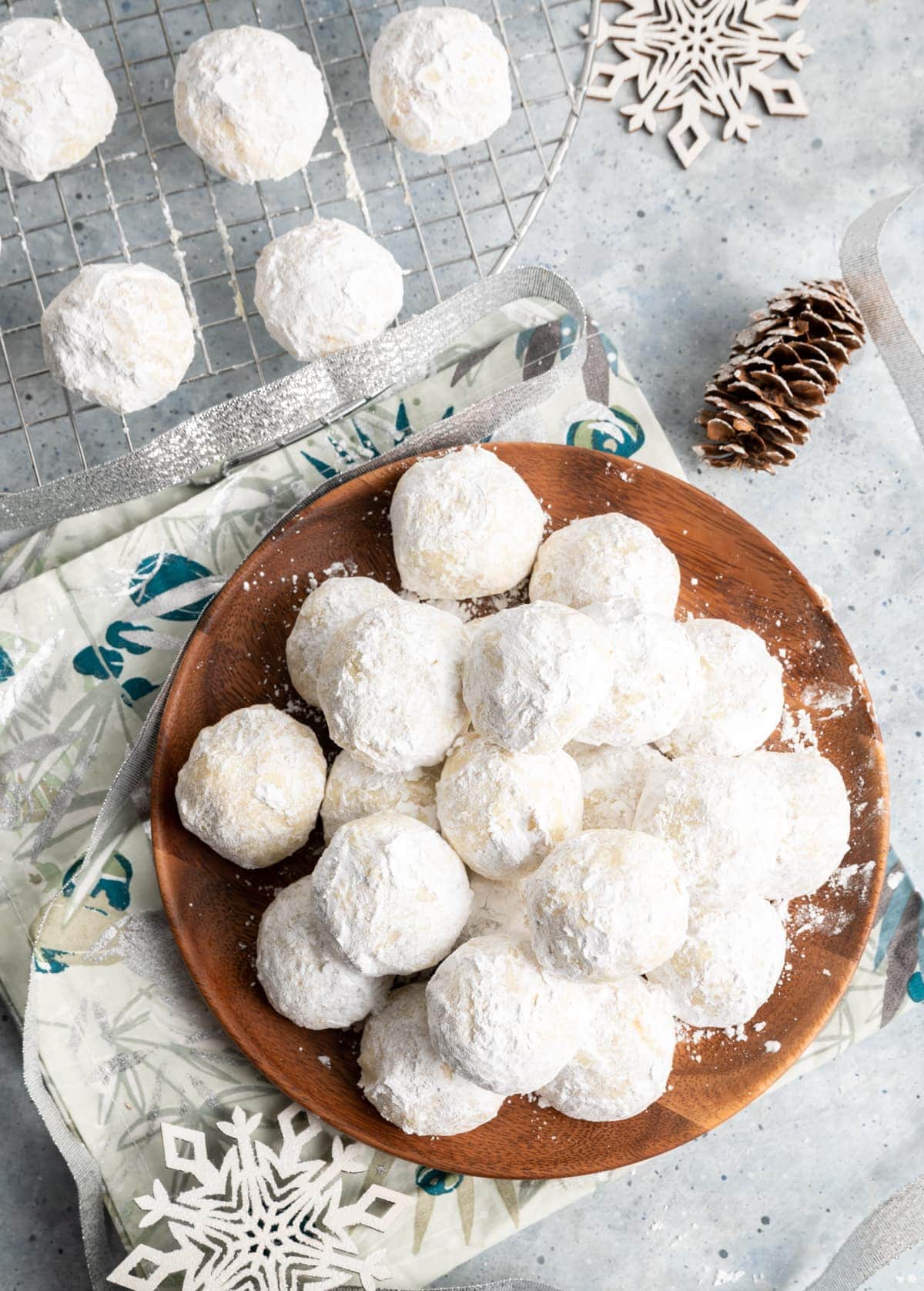 round cookies covered in powdered sugar on a brown plate, wire rack with same cookies, napkin, wood snowflakes, pine cone