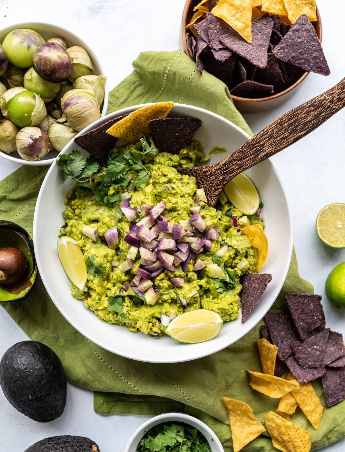 white bowl of tomatillo guacamole with green avocados and purple tomatillos lime slices blue and yellow chips