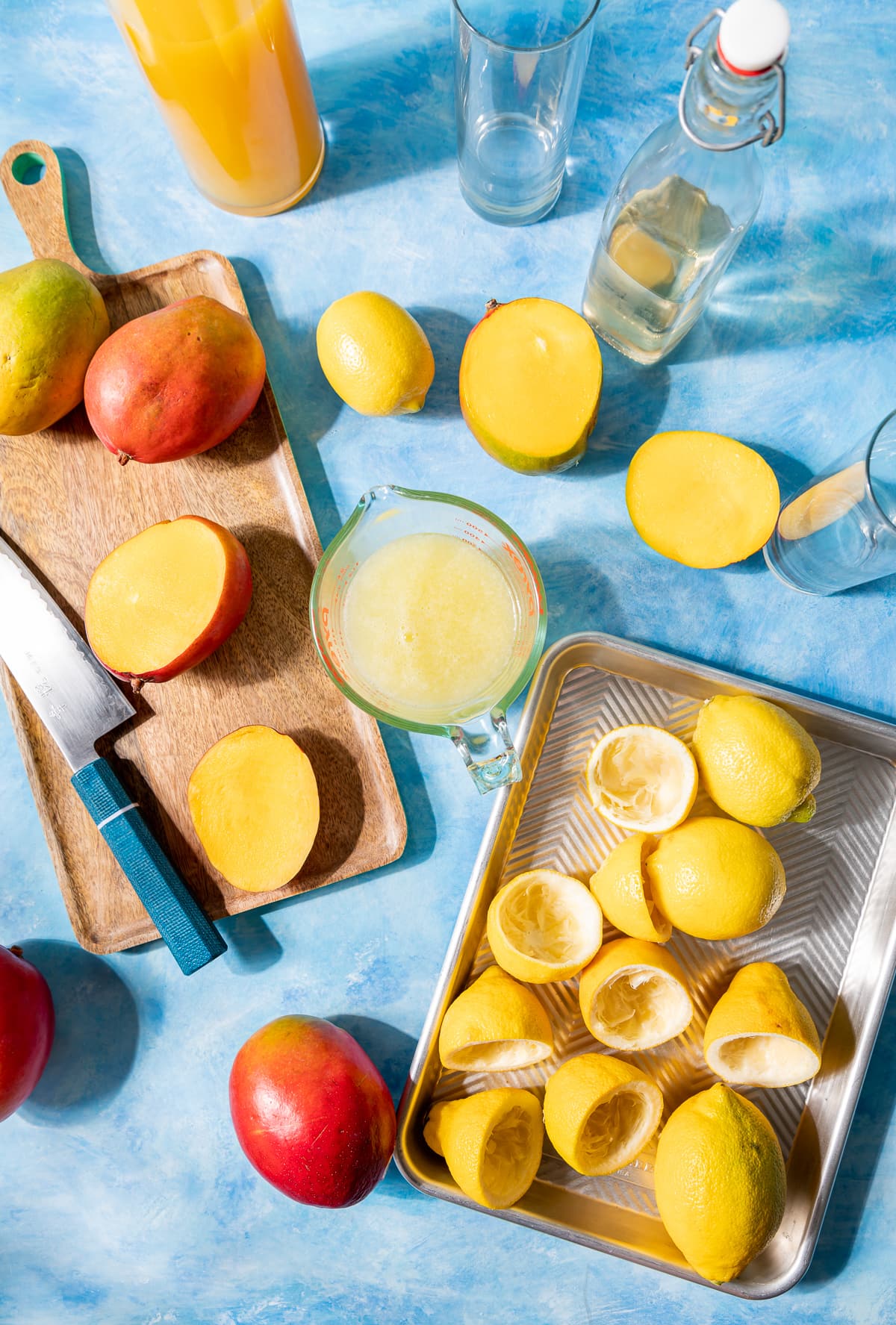 slices of lemon and mangoes glass of lemon juice knife with blue handle whole mangoes whole lemons on silver tray and wood board