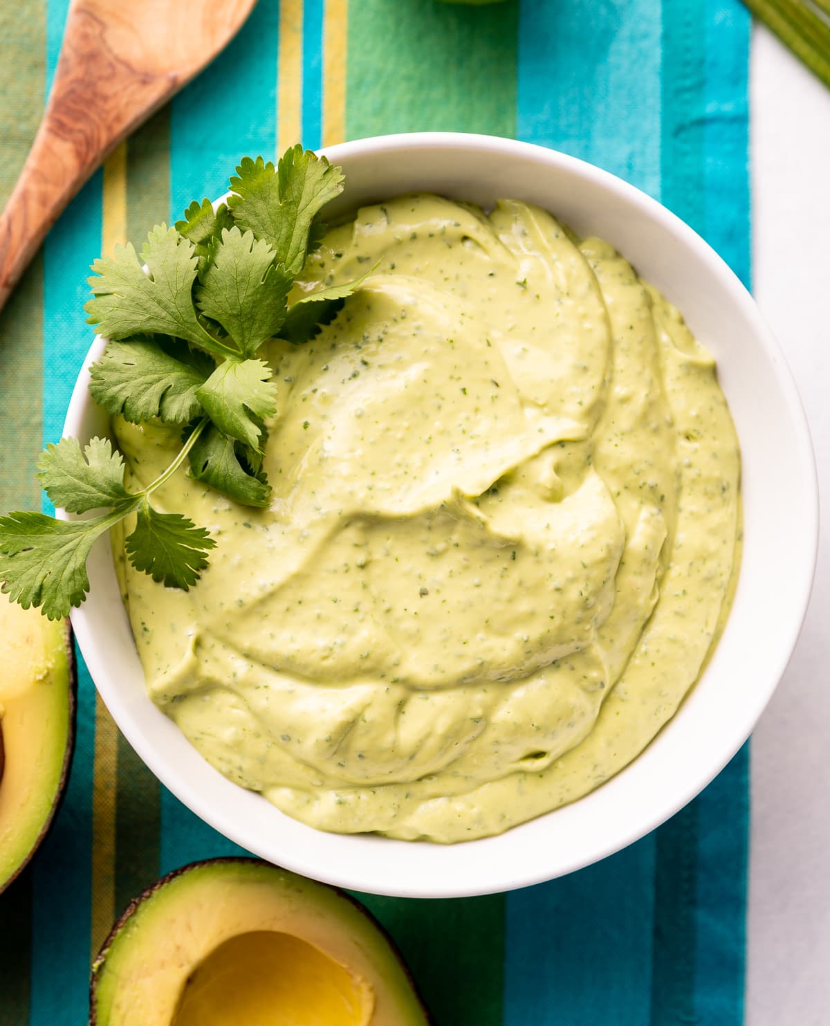 green lime avocado crema sauce in a white bowl with fresh green cilantro avocado cut in half blue striped napkin