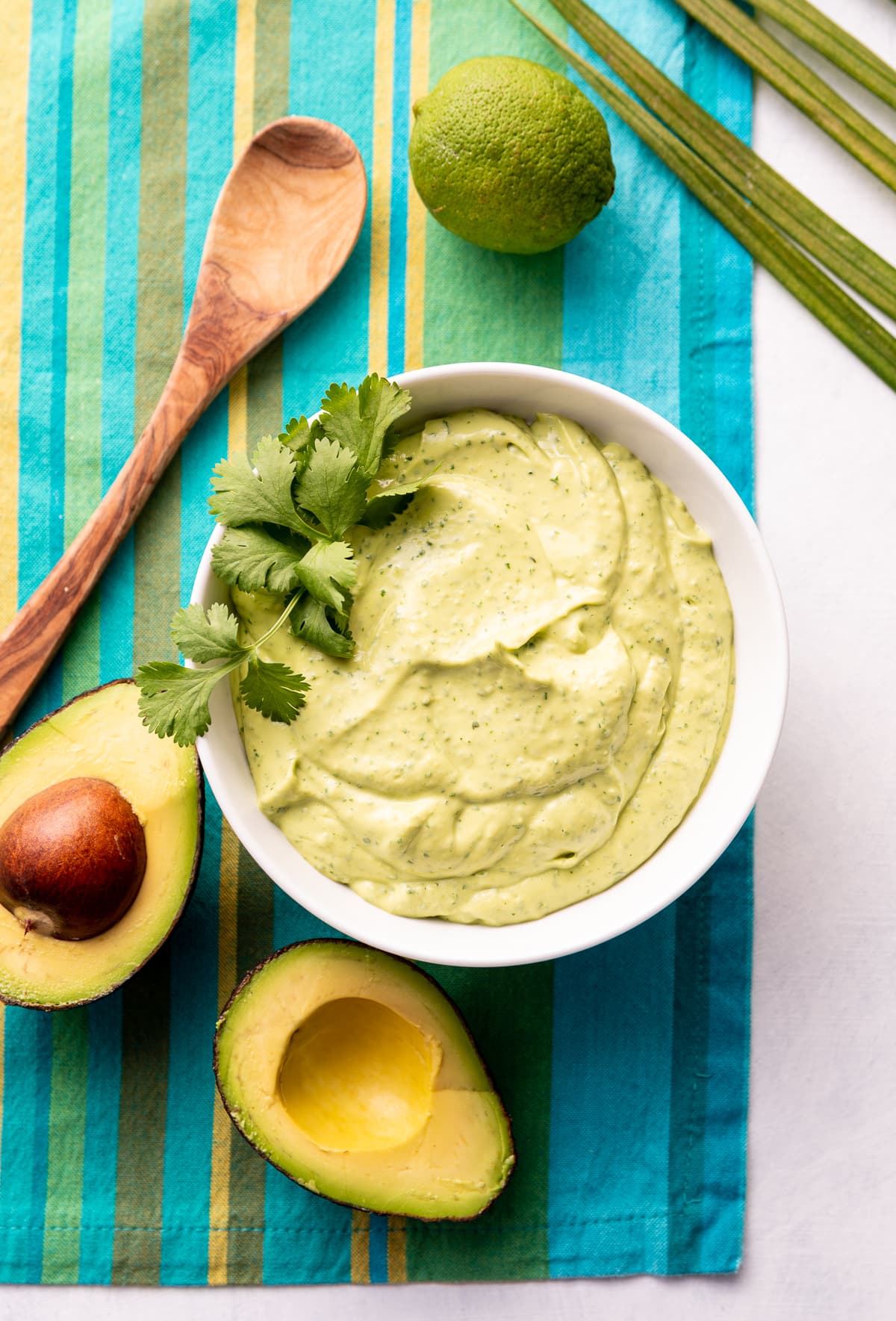 green lime avocado crema sauce in a white bowl with fresh green cilantro avocado cut in half whole lime blue striped napkin