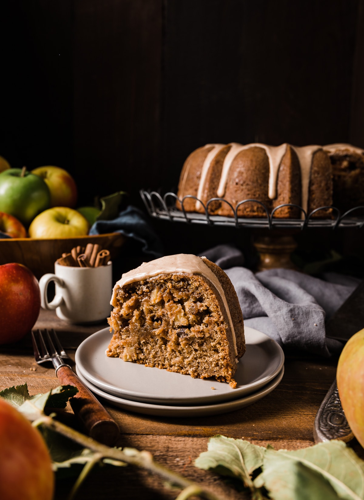 slice of brown sugar bundt cake on a grey cake plate with whole cake in back on a wire cooling rack bowl of apples bowl of cinnamon sticks