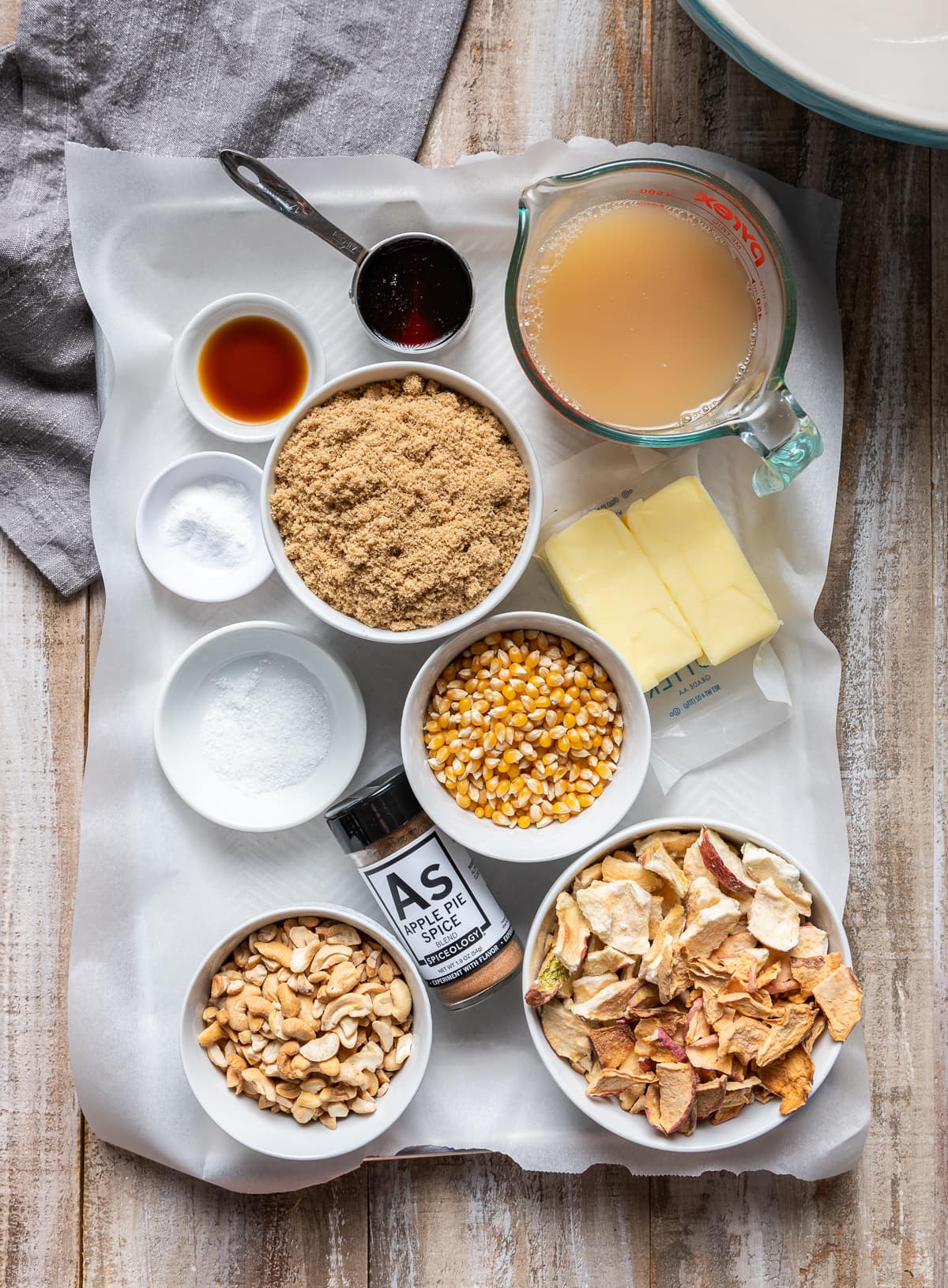 ingredients for caramel apple popcorn on a paper lined tray glass of apple cider bowl of brown sugar butter sticks bowl of cashews bowl of dried apple chips bowl of popcorn kernels bowl of salt bowl of vanilla extract bowl of baking soda