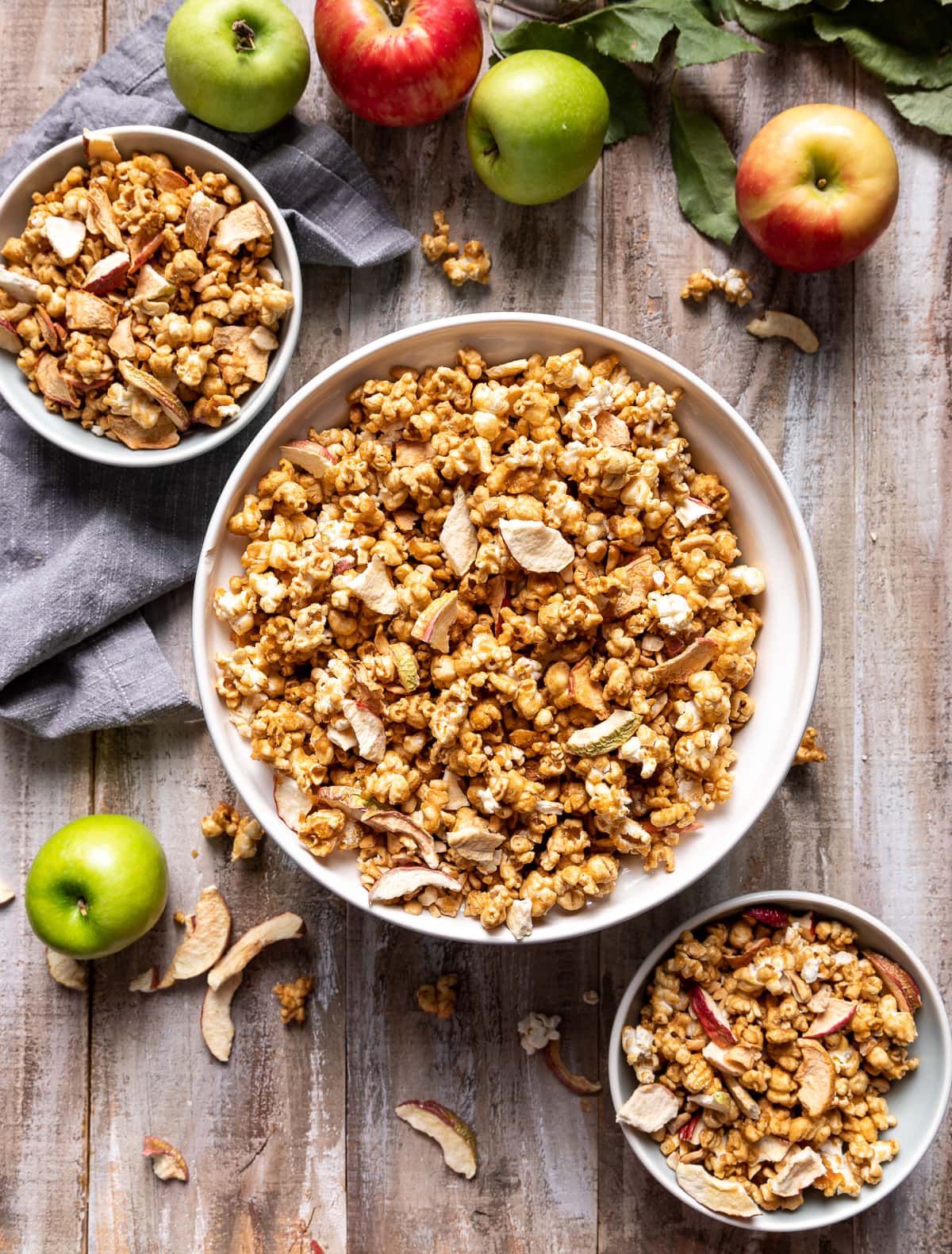 large white bowl and two smaller pale blue bowls filled with brown caramel popcorn pale apple pieces whole green apples whole red apples grey napkin 