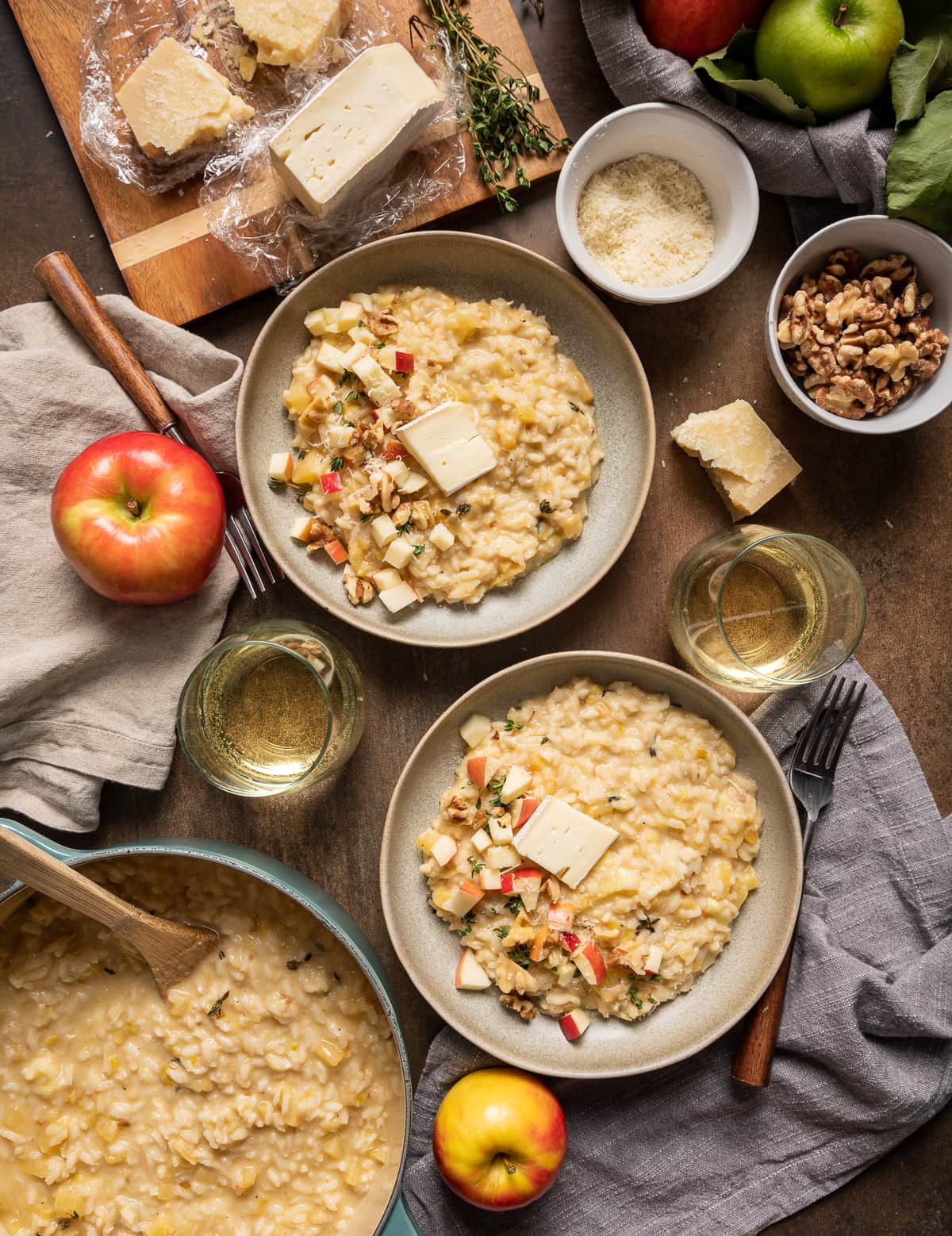 bowls of brie risotto with apples and walnuts glasses of wine pot of brie risotto whole apples cutting board with brie and parmesan cheese