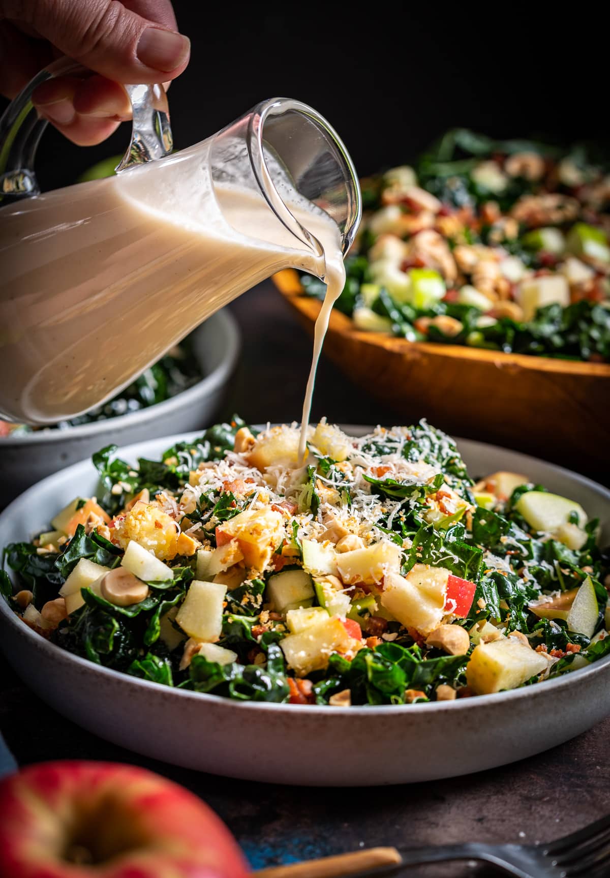 cream colored vinaigrette being poured from a glass bottle over a kale salad with apples hazelnuts shredded parmesan cheese