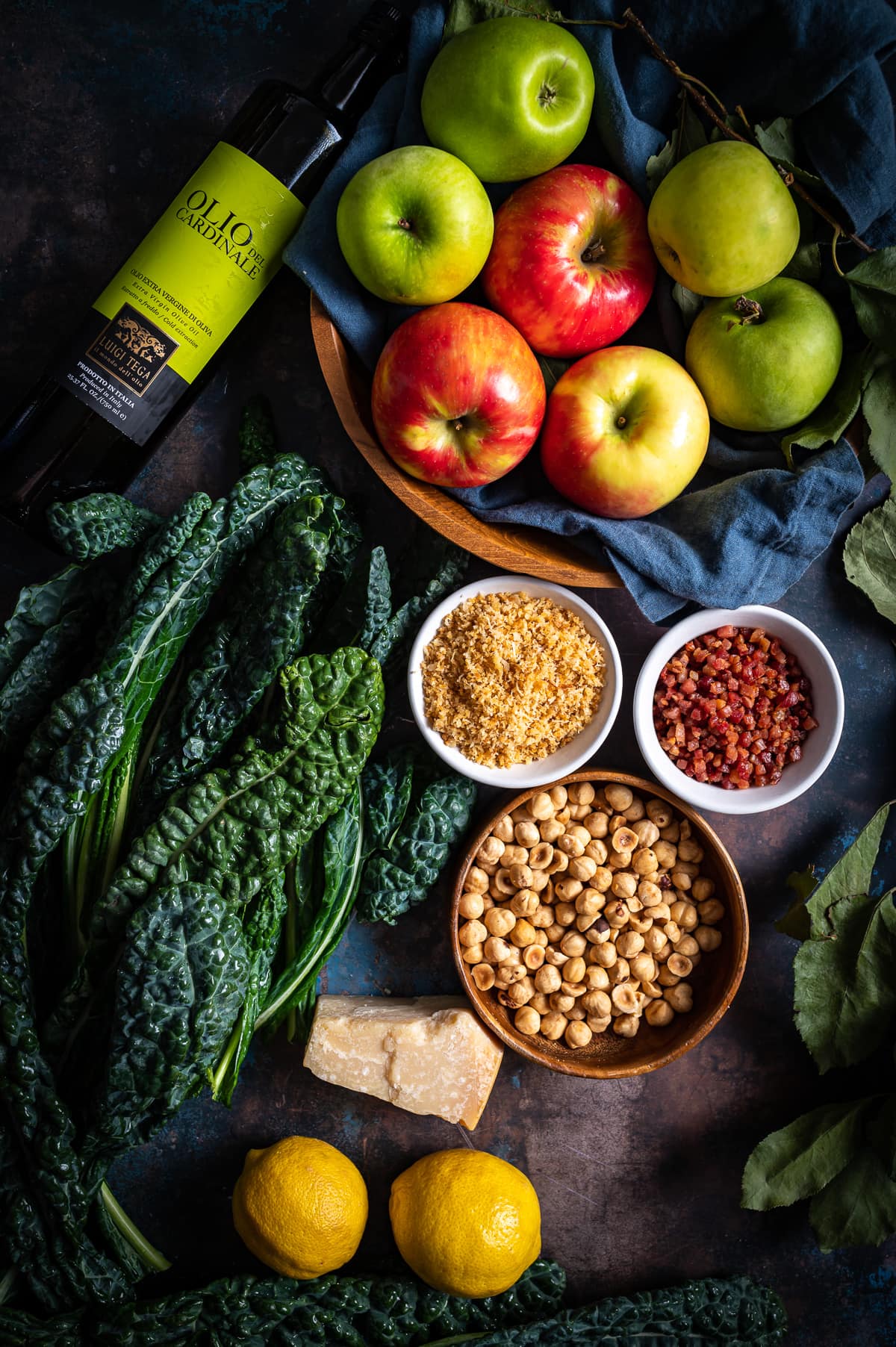 chopped kale salad ingredients bowl of green and red apples bowl of hazelnuts bowl of panko bowl of cooked pancetta lemons tuscan kale bottle of luigi tega oil