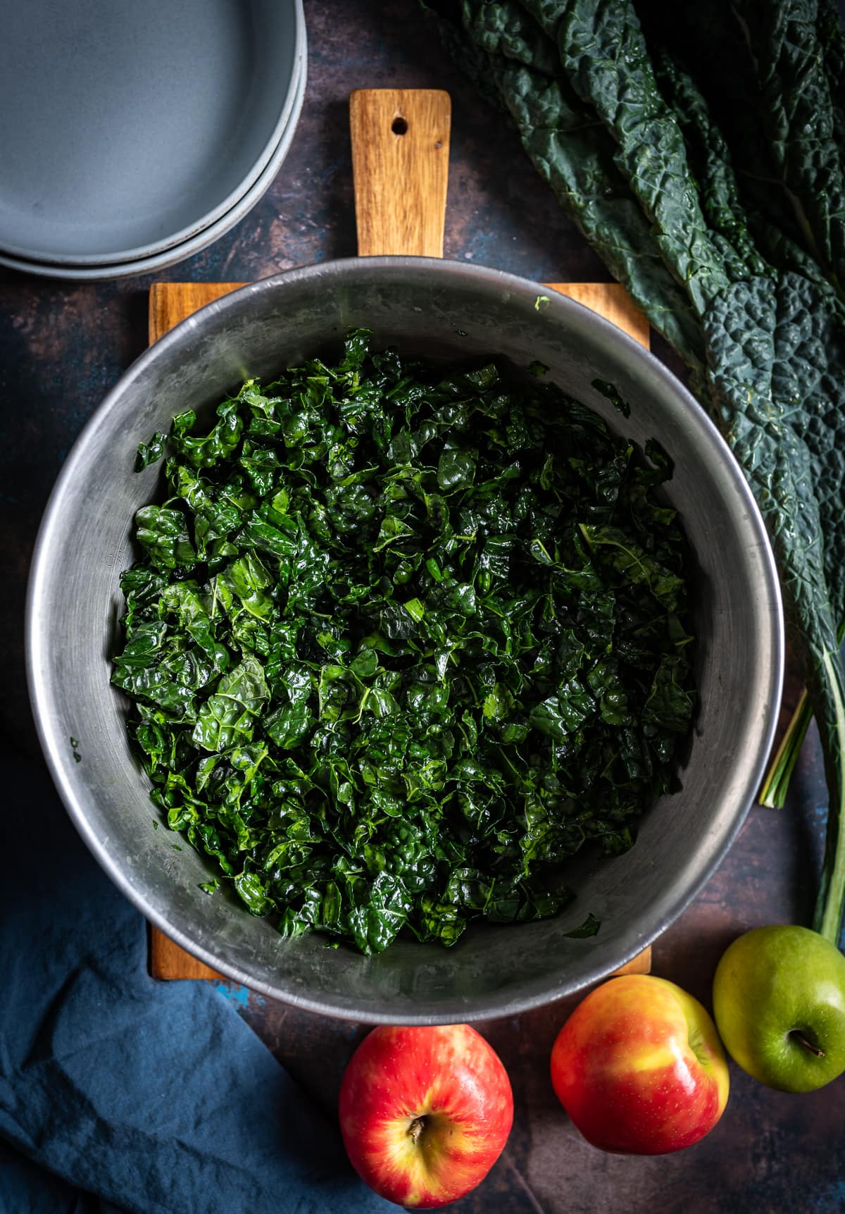 large metal bowl filled with chopped tuscan kale whole red and green apples blue plates