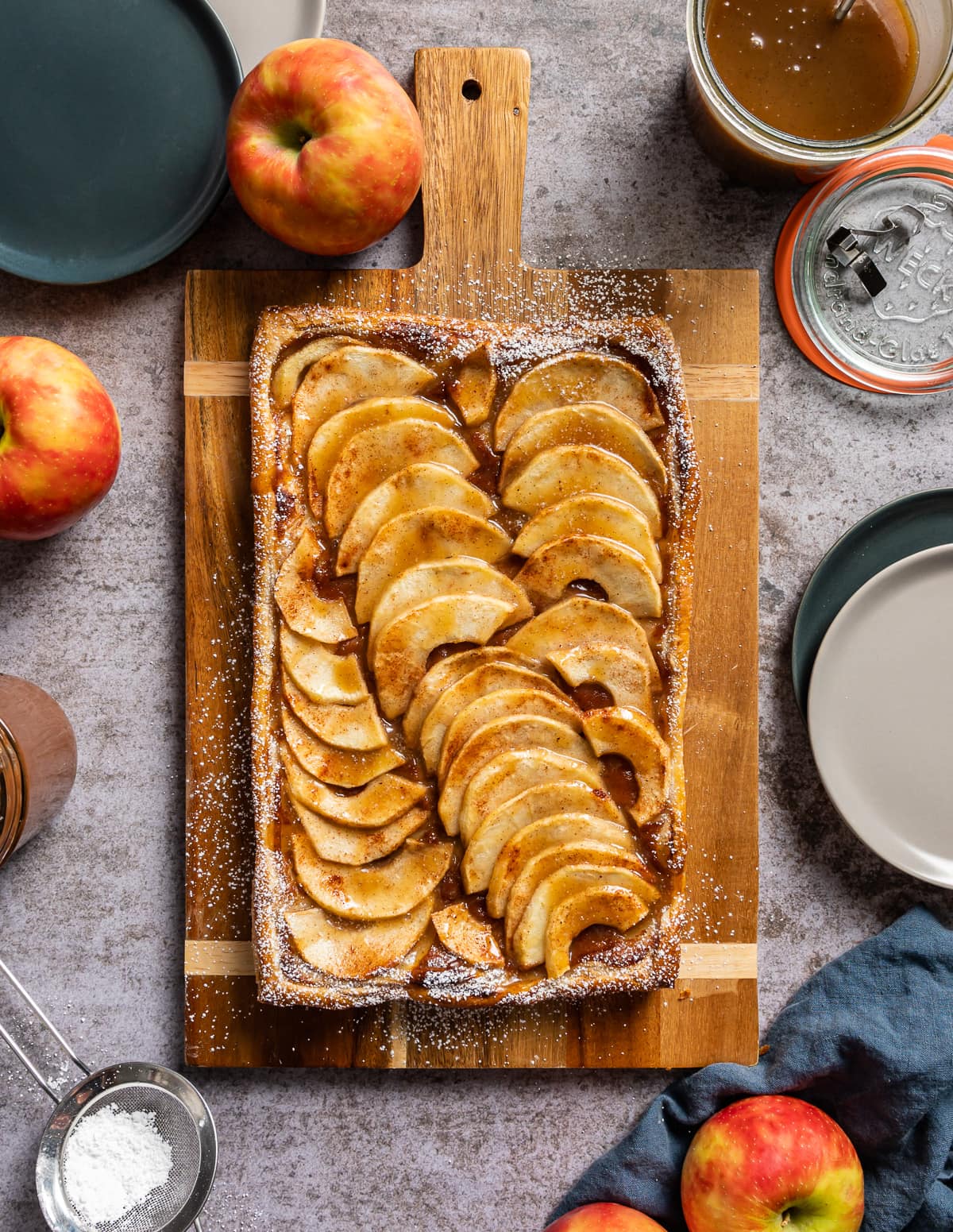 puff pastry apple tart on a cutting board whole red apples strainer with powdered sugar jar of caramel sauce