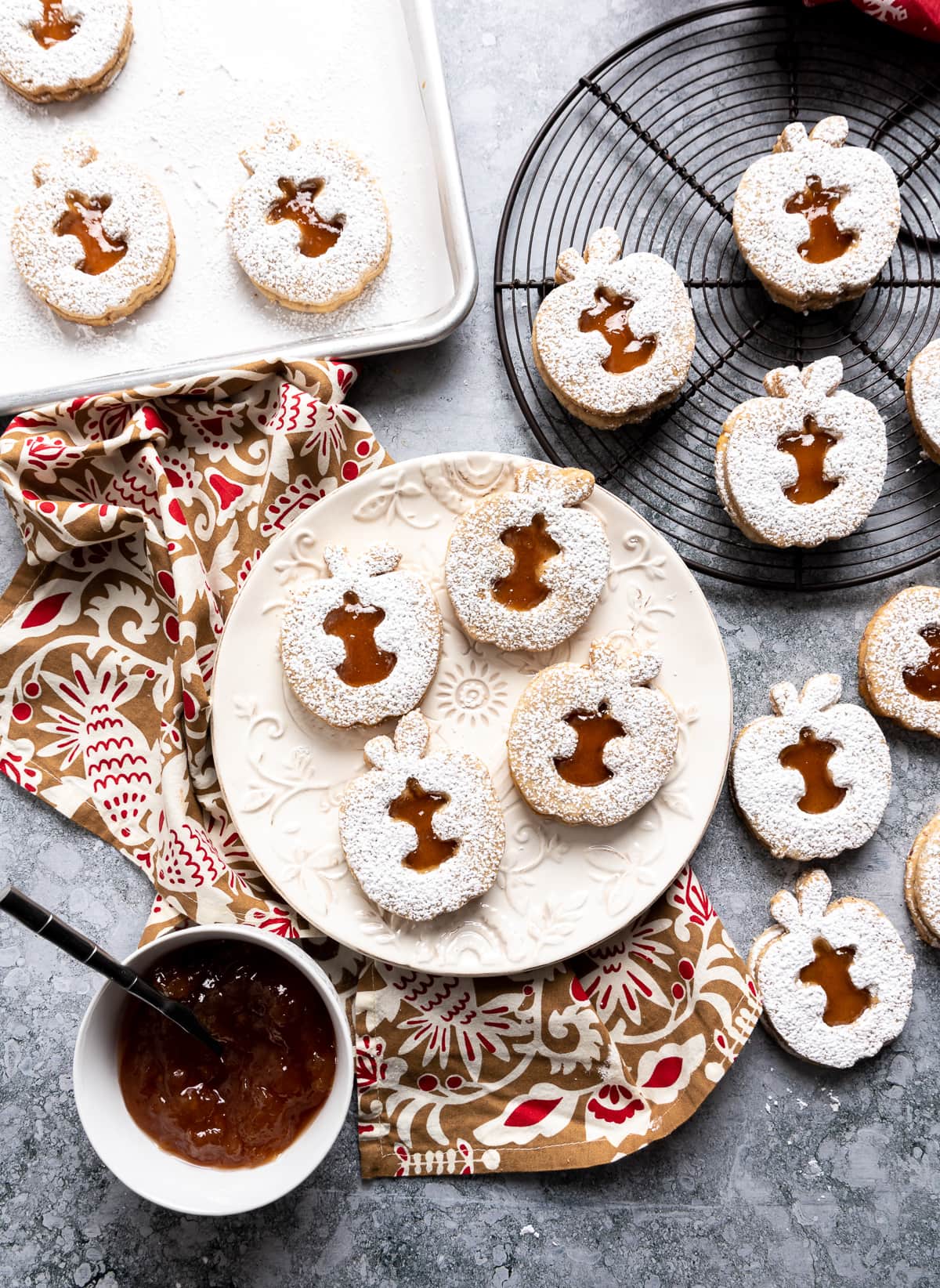 apple shaped linzer cookies with brown apple jam filling on white plate bowl of jam cookies on wire rack