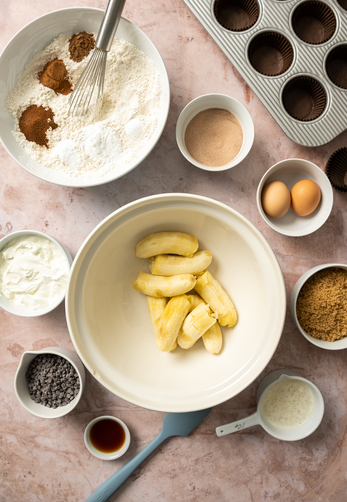 bananas in a bowl bowl of flour and spices bowl of two eggs bowl of sour cream bowl of brown sugar bowl of chocolate chips gray spatula muffin pan with brown liners small bowl of vanilla extract measuring cup with melted butter