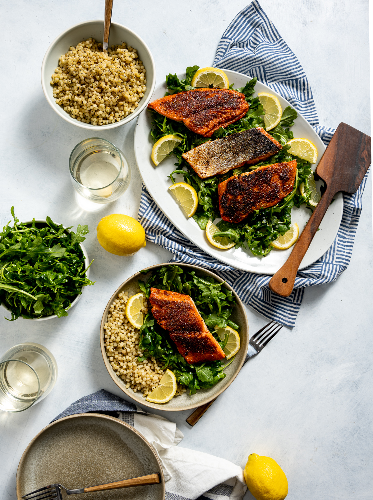 seared sockeye salmon on white and tan plates with green arugula yellow lemon slices bowl of israeli couscous