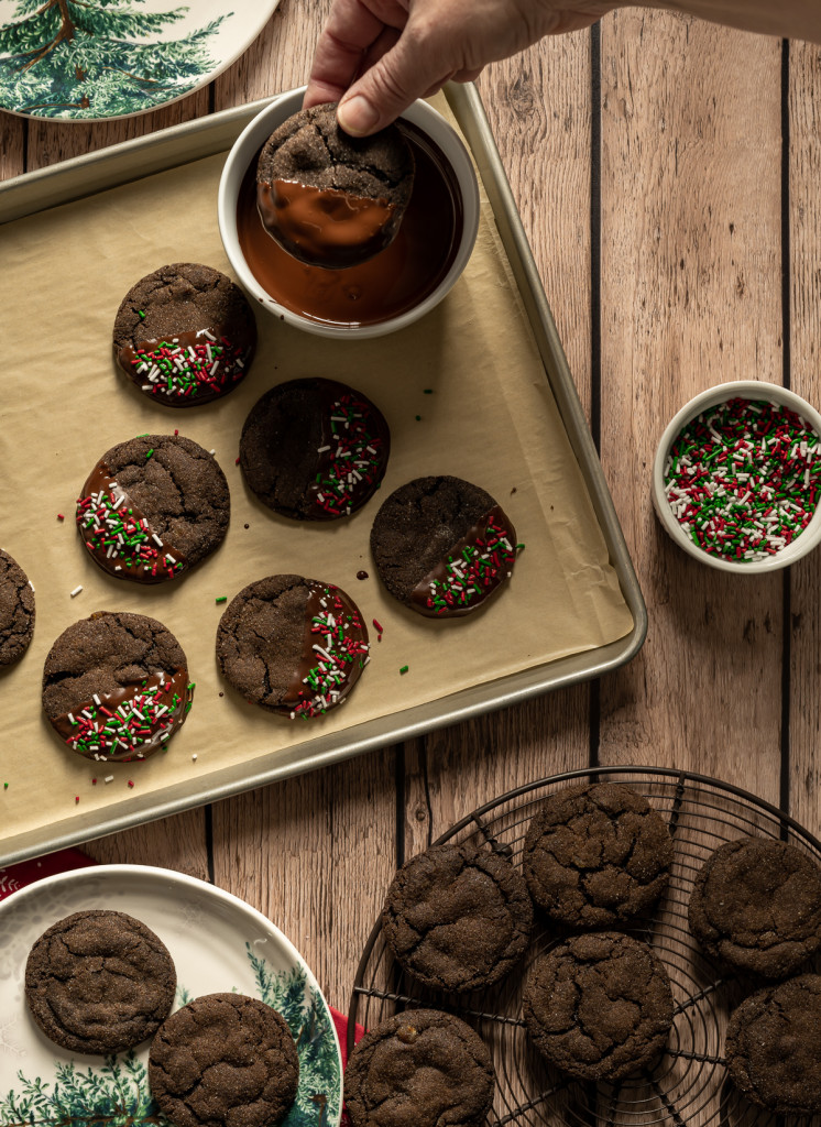 chocolate gingerbread cookies with red green and white sprinkles on a baking tray some cookies without sprinkles on a round wire tray one cookie dipped in chocolate