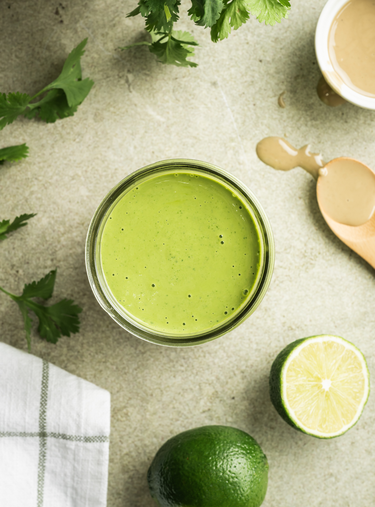 green cilantro lime tahini sauce in a jar limes cilantro wooden spoon small white bowl with tahini green and white checkered cloth