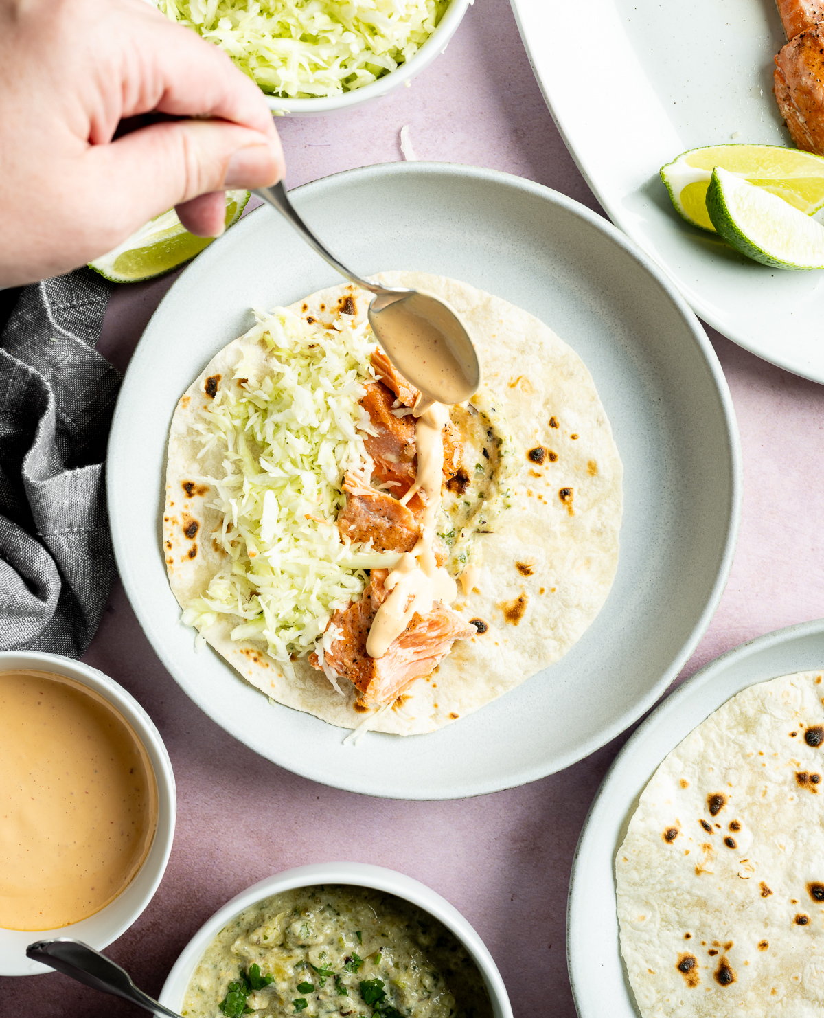 sauce being spooned over salmon green salsa and green shredded cabbage on a tortilla on a light blue plate