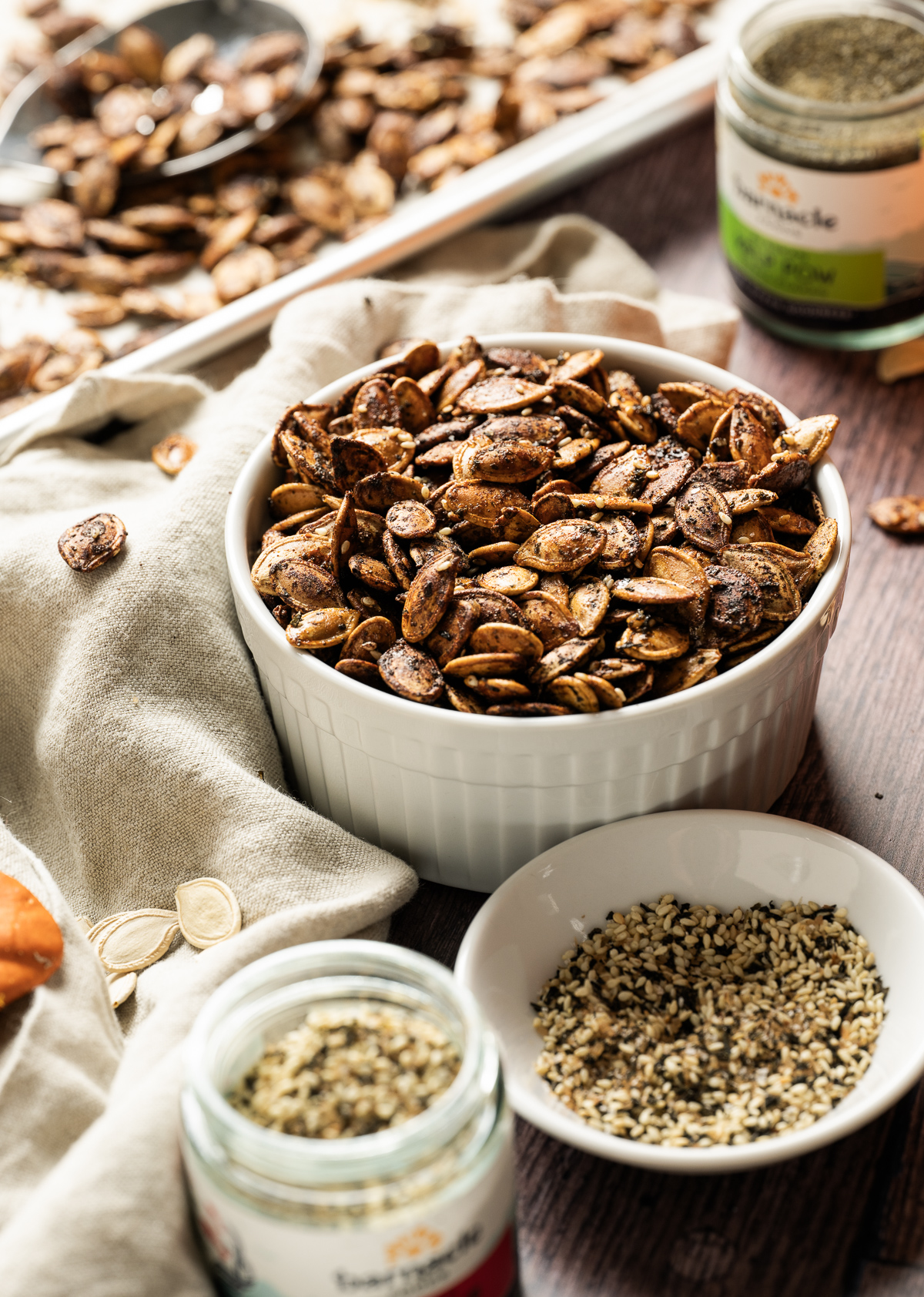 brown roasted pumpkin seeds in white bowl tray of pumpkin seeds jars of seasoning small bowl of furikake brown napkin