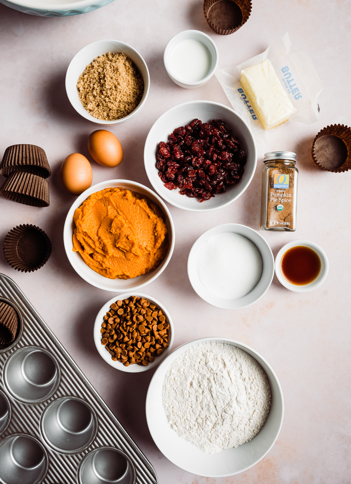 bowl of pumpkin bowl of sugar bowl of cranberries stick of butter paper muffin liners eggs bowl of flour bowl of cinnamon chips jar of pumpkin pie spice