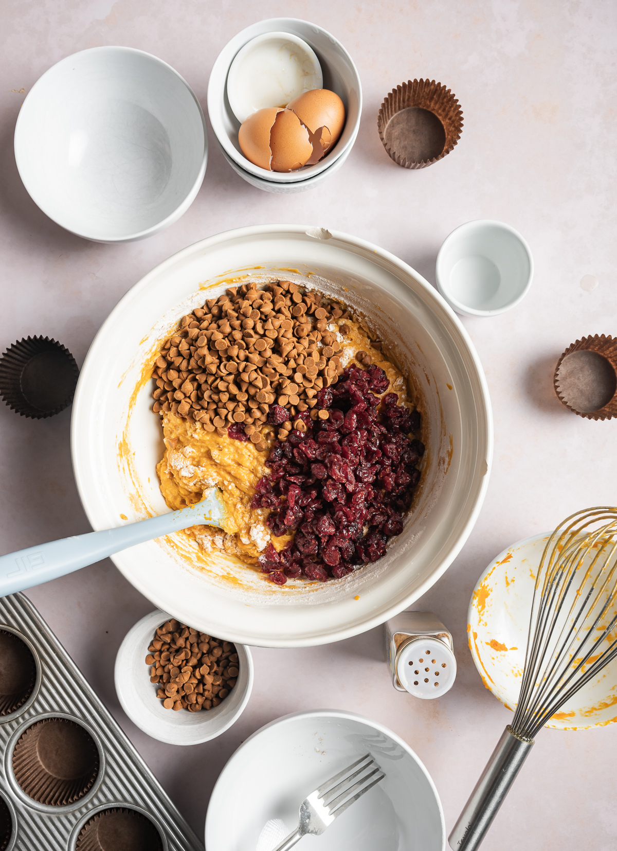 cinnamon chips and dried cranberry pieces on top of pumpkin muffin batter muffin in a bowl, various bowls of separate muffin ingredients