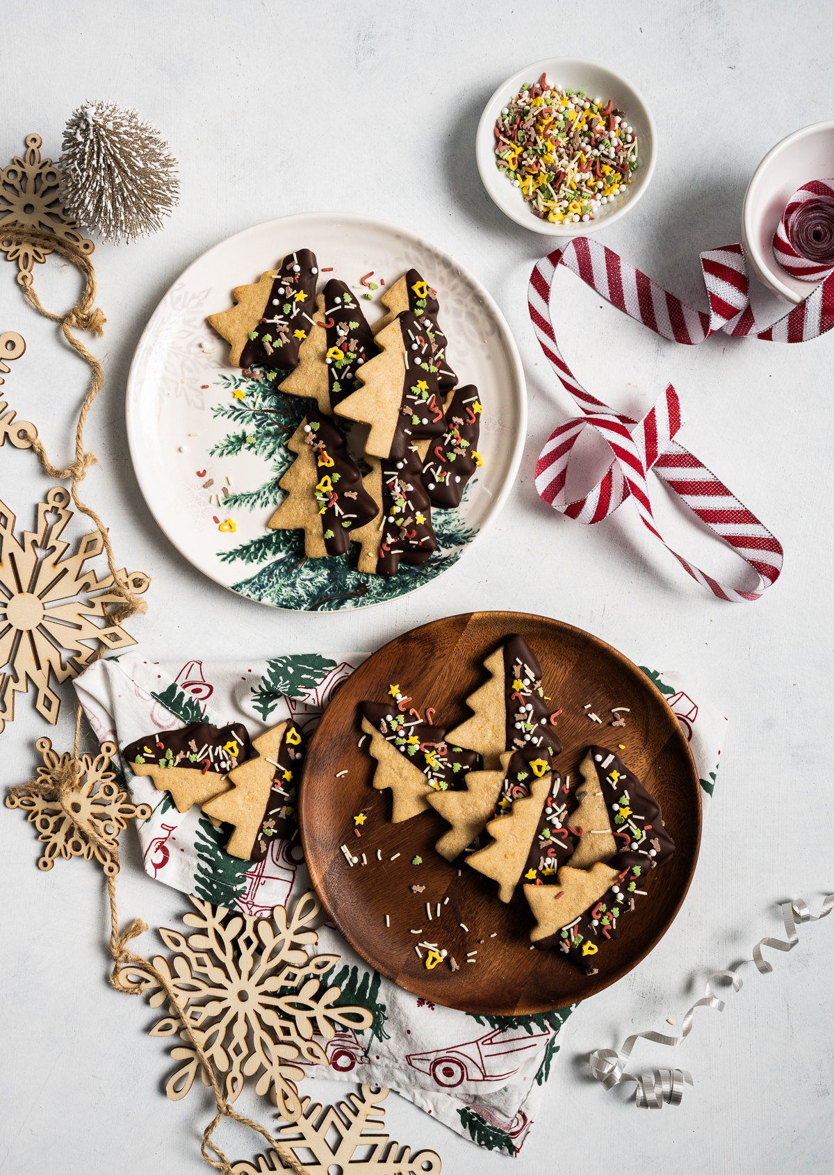 tree shaped cinnamon shortbread cookies half dipped in chocolate with sprinkles bowl of sprinkles ornaments red and white striped ribbon