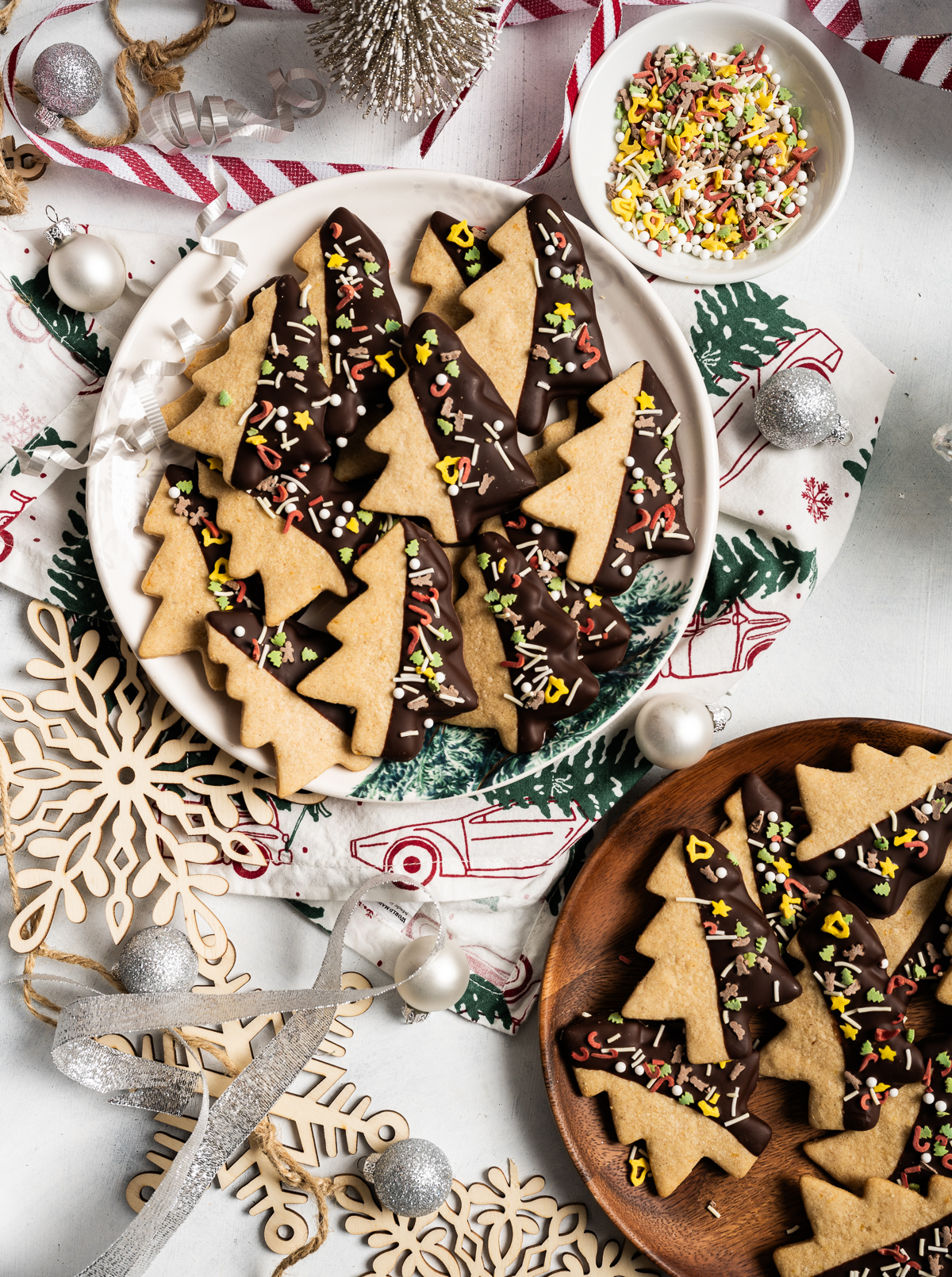 tree shaped cinnamon shortbread cookies half dipped in chocolate with sprinkles bowl of sprinkles ornaments red and white striped ribbon